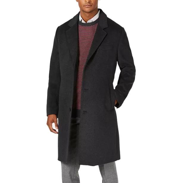 A man is wearing a long, dark overcoat with a classic notch lapel, layered over a patterned sweater and collared shirt. The coat features a button-up front and side pockets, paired with light gray trousers.