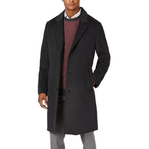 A man is wearing a long, dark overcoat with a classic notch lapel, layered over a patterned sweater and collared shirt. The coat features a button-up front and side pockets, paired with light gray trousers.