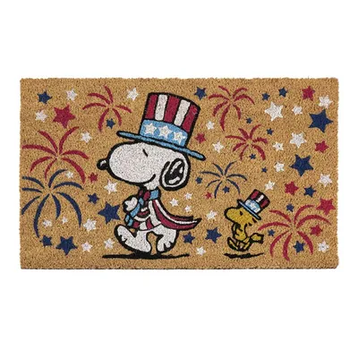 A doormat featuring Snoopy and Woodstock wearing patriotic hats and scarves, surrounded by red, white, and blue stars and fireworks on a tan background.