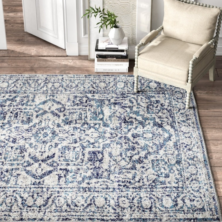 A patterned area rug with blue and white hues lies on wooden flooring beside a beige upholstered chair with spindle detailing. A stack of books and a vase with greenery are nearby.
