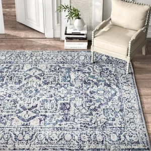 A patterned area rug with blue and white hues lies on wooden flooring beside a beige upholstered chair with spindle detailing. A stack of books and a vase with greenery are nearby.