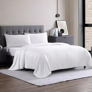 White queen-sized comforter on a neatly made bed with a gray tufted headboard; part of a modern, minimalistic bedroom setup featuring a gray nightstand, decorative books, and a framed artwork, all on a soft, light-colored rug.