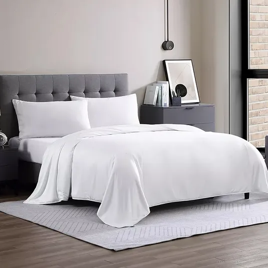 White queen-sized comforter on a neatly made bed with a gray tufted headboard; part of a modern, minimalistic bedroom setup featuring a gray nightstand, decorative books, and a framed artwork, all on a soft, light-colored rug.