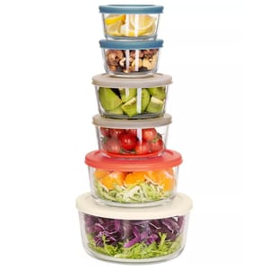 A 12-piece glass bowl set featuring six stackable round bowls with multi-colored plastic lids. These bowls contain an assortment of fruits, vegetables, and salads, demonstrating their use for food storage or serving.