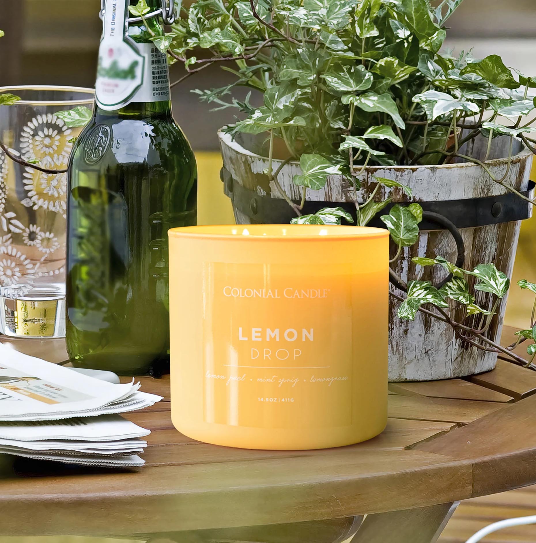 Yellow Colonial Candle with \“Lemon Drop\“ scent, featuring lemon peel and lemongrass aromas, next to a green glass bottle, a decorative drinking glass, and a potted plant on a wooden table.