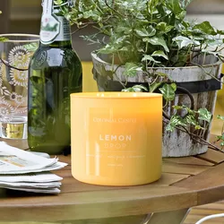 Yellow Colonial Candle with \“Lemon Drop\“ scent, featuring lemon peel and lemongrass aromas, next to a green glass bottle, a decorative drinking glass, and a potted plant on a wooden table.