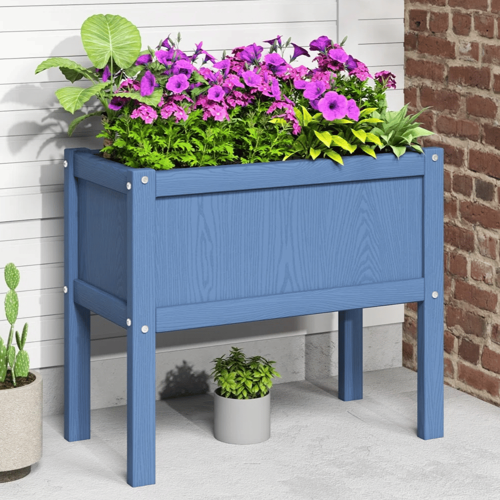 A blue wooden raised planter box filled with green and purple flowers is against a brick and white siding backdrop. Nearby, a potted cactus and a small plant in a gray pot add decorative touches.