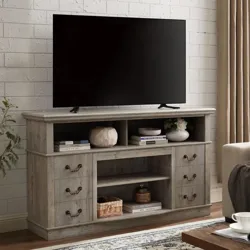 A wooden TV stand with drawers and shelves holds a flat-screen TV. Decorations, books, and a woven basket are arranged on the shelves, complemented by a cream curtain and a patterned rug.