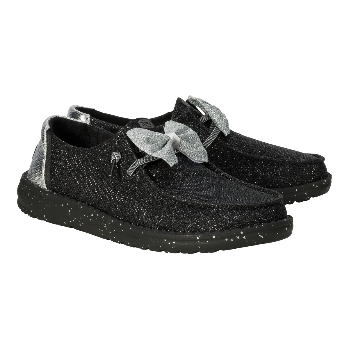 Hey Dude Wendy Sparkle Bow shoes feature a black, glittery finish with a large silver bow on the top. They have a speckled black sole and metallic silver detailing at the heel, offering a stylish and casual look.
