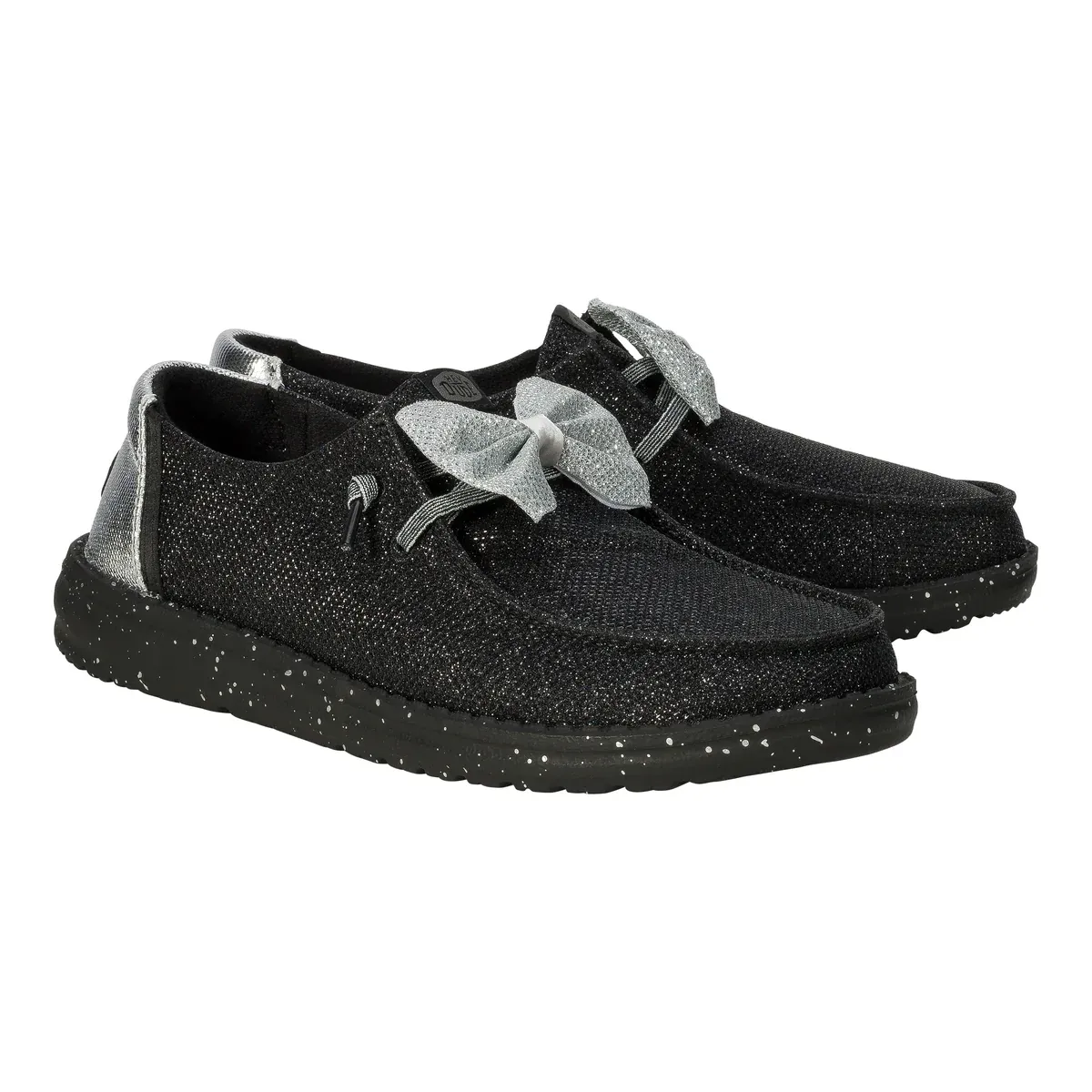 Hey Dude Wendy Sparkle Bow shoes feature a black, glittery finish with a large silver bow on the top. They have a speckled black sole and metallic silver detailing at the heel, offering a stylish and casual look.