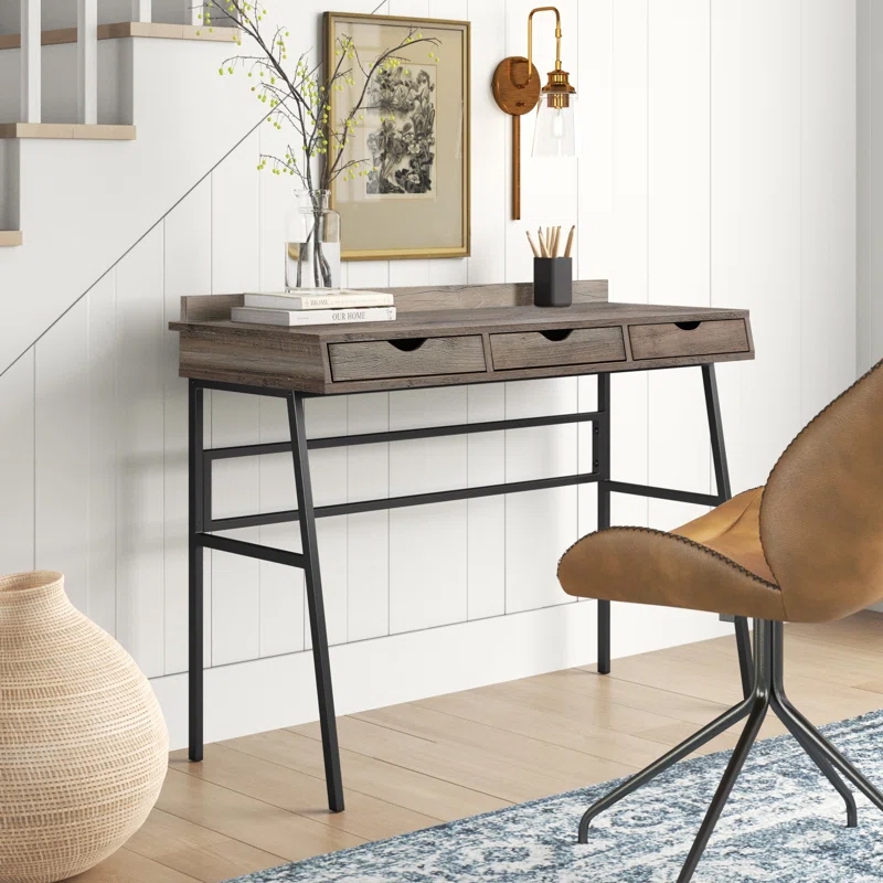 A modern desk with a wood finish and black metal legs is paired with a leather swivel chair. The desk features three small drawers, topped with books and a decorative vase.