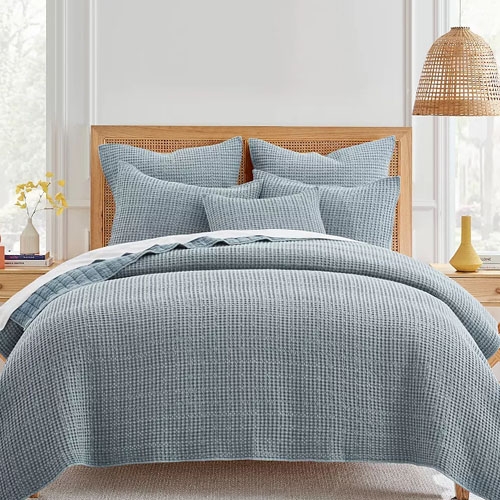 A blue textured comforter set with pillows on a bed, framed by a woven headboard and a wicker pendant lamp overhead.