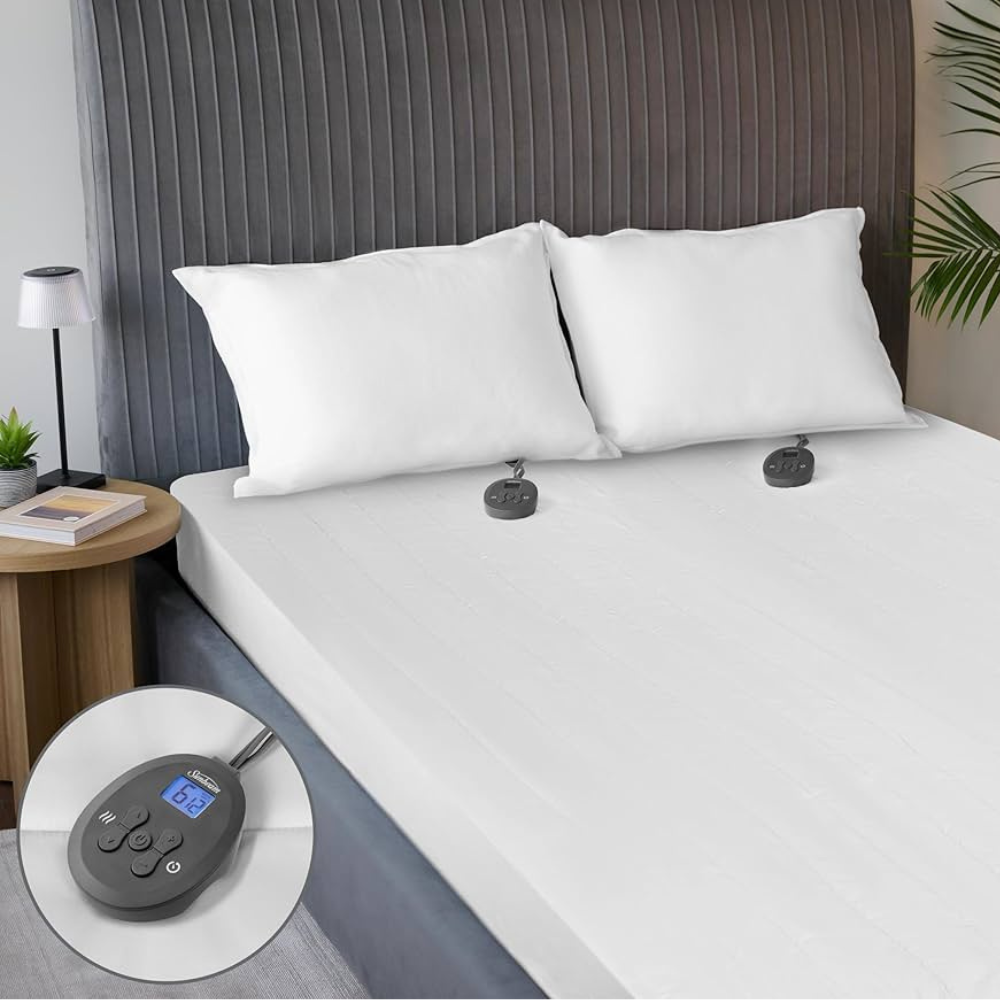 An electric heated mattress pad is on a bed with a grey headboard, featuring dual controllers shown in close-up. Two white pillows are placed on the bed, and a small side table with a lamp and plant is beside it.