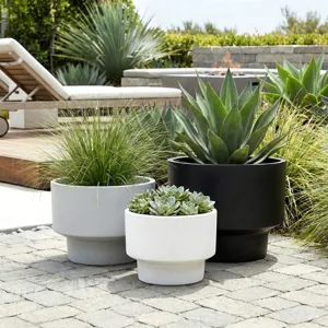 Three modern planters are shown: a black one containing an agave plant, a grey one with ornamental grass, and a white one with succulents, set on a patio with a lounge chair and fire pit in the background.