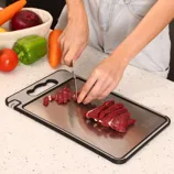 A person is cutting raw meat on a metal cutting board surrounded by vegetables including a green pepper, carrot, onion, and tomato.