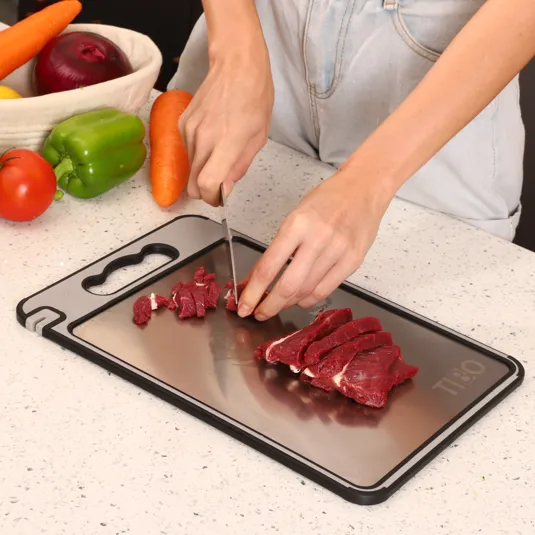 A person is cutting raw meat on a metal cutting board surrounded by vegetables including a green pepper, carrot, onion, and tomato.
