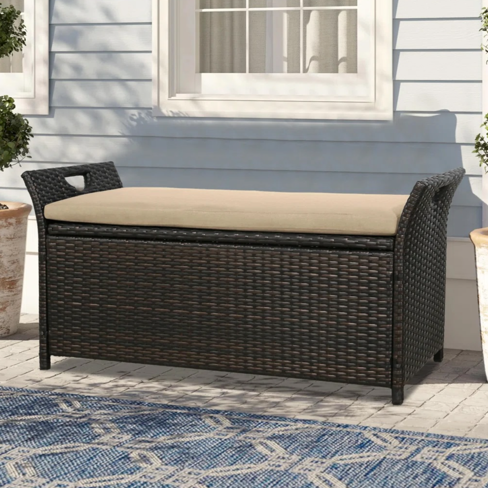 Outdoor storage bench with a dark wicker finish and beige cushion, flanked by potted plants and placed on a patterned blue rug in front of a light blue wall.