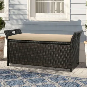 Outdoor storage bench with a dark wicker finish and beige cushion, flanked by potted plants and placed on a patterned blue rug in front of a light blue wall.