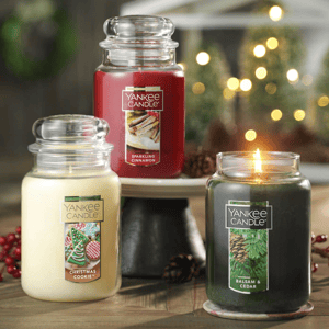 Three Yankee Candles: \“Sparkling Cinnamon\“ in red, \“Christmas Cookie\“ in white, and a lit \“Balsam & Cedar\“ in green, set against a festive background with bokeh lights.