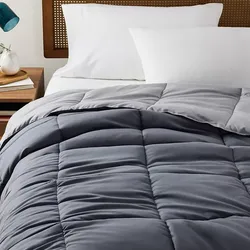 A gray quilted comforter is on a bed with white pillows and a wooden headboard. A wooden nightstand with a vase and books is beside the bed.