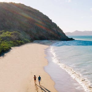 A couple walks along a pristine, sandy beach next to clear blue ocean water, with lush green hills and a scenic mountain view in the background.