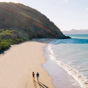 A couple walks along a pristine, sandy beach next to clear blue ocean water, with lush green hills and a scenic mountain view in the background.