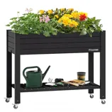 A raised garden bed on wheels with a shelf below, filled with various flowering plants. Gardening tools, a watering can, and plant pots are placed on the lower shelf.
