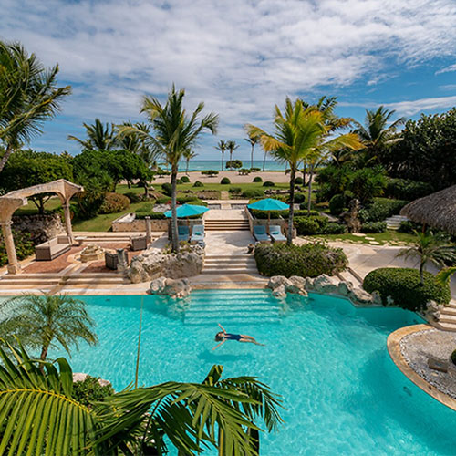 A luxurious tropical resort features a swimming pool surrounded by palm trees, lounge chairs with blue umbrellas, and a view of the ocean in the background.