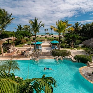 A luxurious tropical resort features a swimming pool surrounded by palm trees, lounge chairs with blue umbrellas, and a view of the ocean in the background.