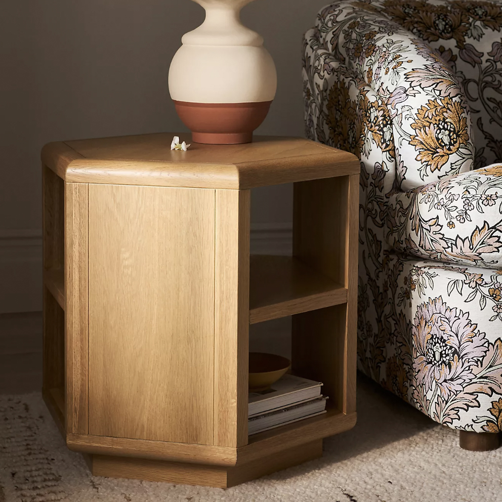 Wooden hexagonal side table with open shelves next to a floral-patterned sofa, topped with a round vase.