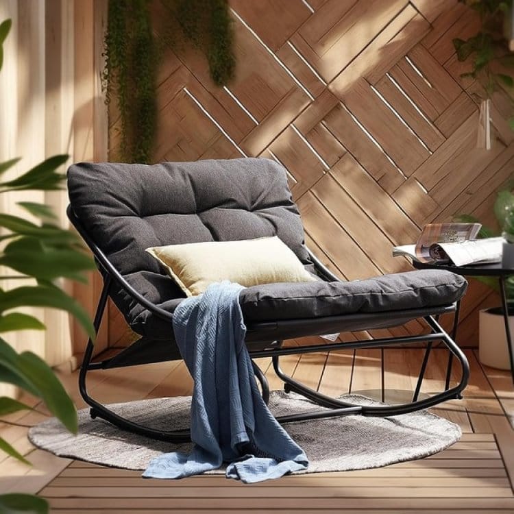 A cushioned papasan chair with a throw blanket and a small pillow on a round rug, near a side table with a book.