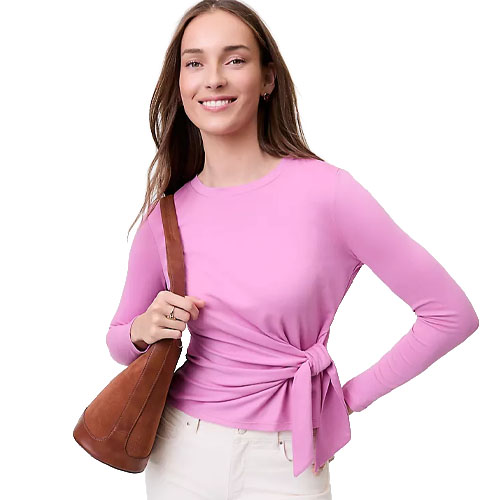 A person in a pink long-sleeve top with a side tie is carrying a brown suede shoulder bag. They are wearing white pants.