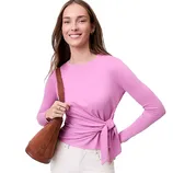A person in a pink long-sleeve top with a side tie is carrying a brown suede shoulder bag. They are wearing white pants.