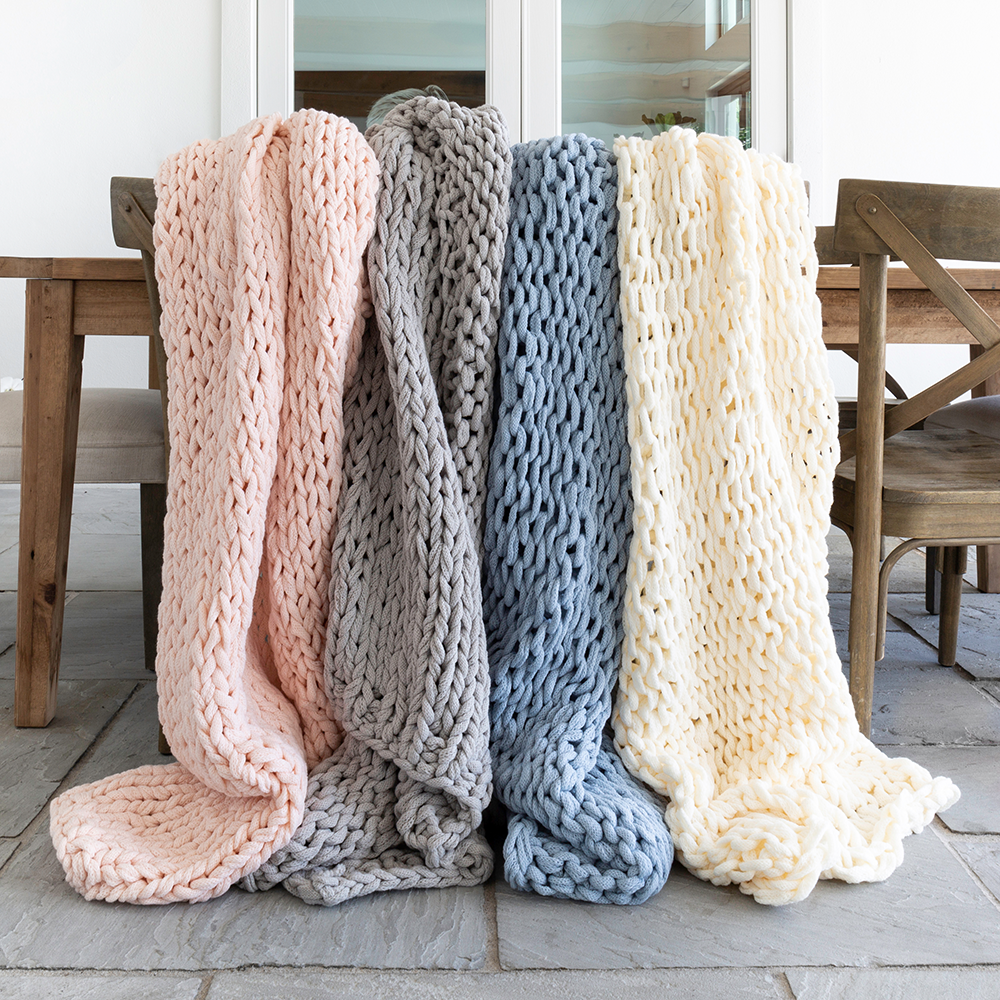 Four chunky knit blankets in pink, gray, blue, and cream draped over a wooden bench.