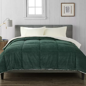 A green corduroy comforter with a sherpa lining covers a bed with cream pillows and a gray headboard. A small wooden side table holds a lamp, next to a framed picture on a white paneled wall.