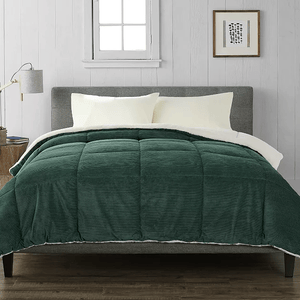 A green corduroy comforter with a sherpa lining covers a bed with cream pillows and a gray headboard. A small wooden side table holds a lamp, next to a framed picture on a white paneled wall.