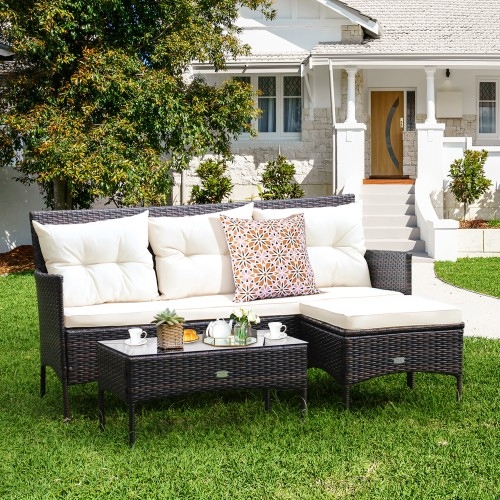 Outdoor wicker patio set with cream cushions and decorative pillows, featuring a sectional sofa and coffee table adorned with tea and cups, set on a grassy lawn in front of a house.