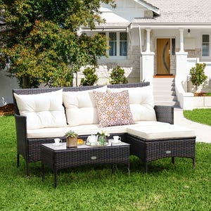 Outdoor wicker patio set with cream cushions and decorative pillows, featuring a sectional sofa and coffee table adorned with tea and cups, set on a grassy lawn in front of a house.