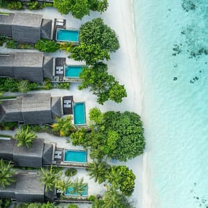 Aerial view of beachfront villas with private pools, surrounded by lush greenery and situated along a sandy shore next to clear turquoise water.