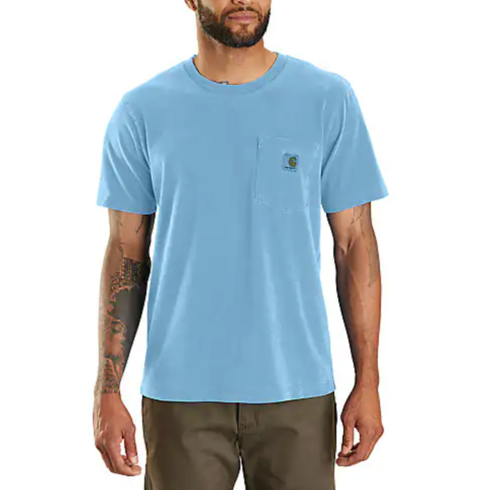 A man is wearing a light blue short-sleeve T-shirt with a chest pocket featuring a small logo, paired with dark brown pants. The T-shirt has a simple, casual design.