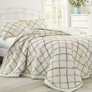 A bed is dressed with a quilt and matching pillowcases, featuring a white background with a colorful, textured grid pattern. A light and airy bedroom setup is visible with a white chest and green plant beside a large window.