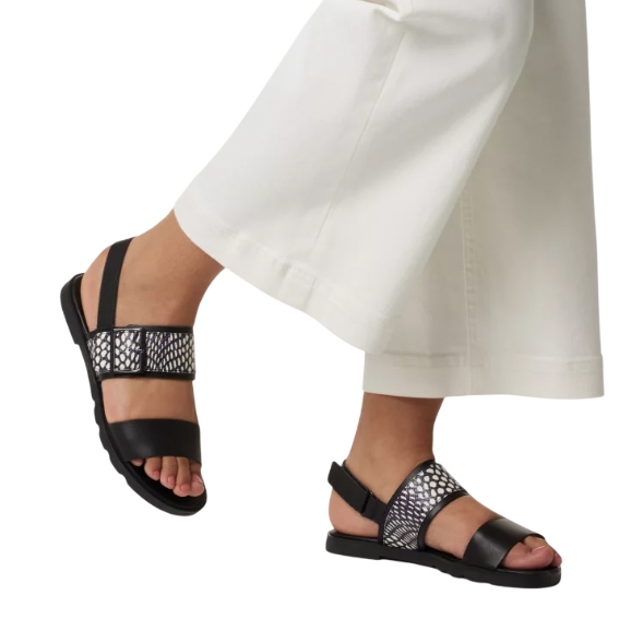 Sorel Slingback Sandals feature black leather straps with a stylish mesh design on the front and an adjustable slingback for a secure fit. They're paired with wide-legged white pants, highlighting the sandals' chic and modern appearance.