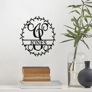 A round, black metal wall decoration features the letter \“J\“ with \“JONES\“ written across it, adorned with a leafy border. It is displayed on a wall beside a clear glass vase with a plant and a stack of books.