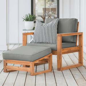 A thick-cushioned wood patio chair set with gray cushions includes a chair and ottoman, featuring a simple wooden frame, complemented by a black and white patterned throw pillow, situated on a wooden deck with a potted plant in the background.
