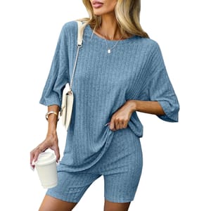 A woman is wearing a blue ribbed knit set, consisting of a loose-fitting top and matching shorts, accessorized with a cream-colored handbag, bracelets, and a necklace. She is holding a white takeaway coffee cup.