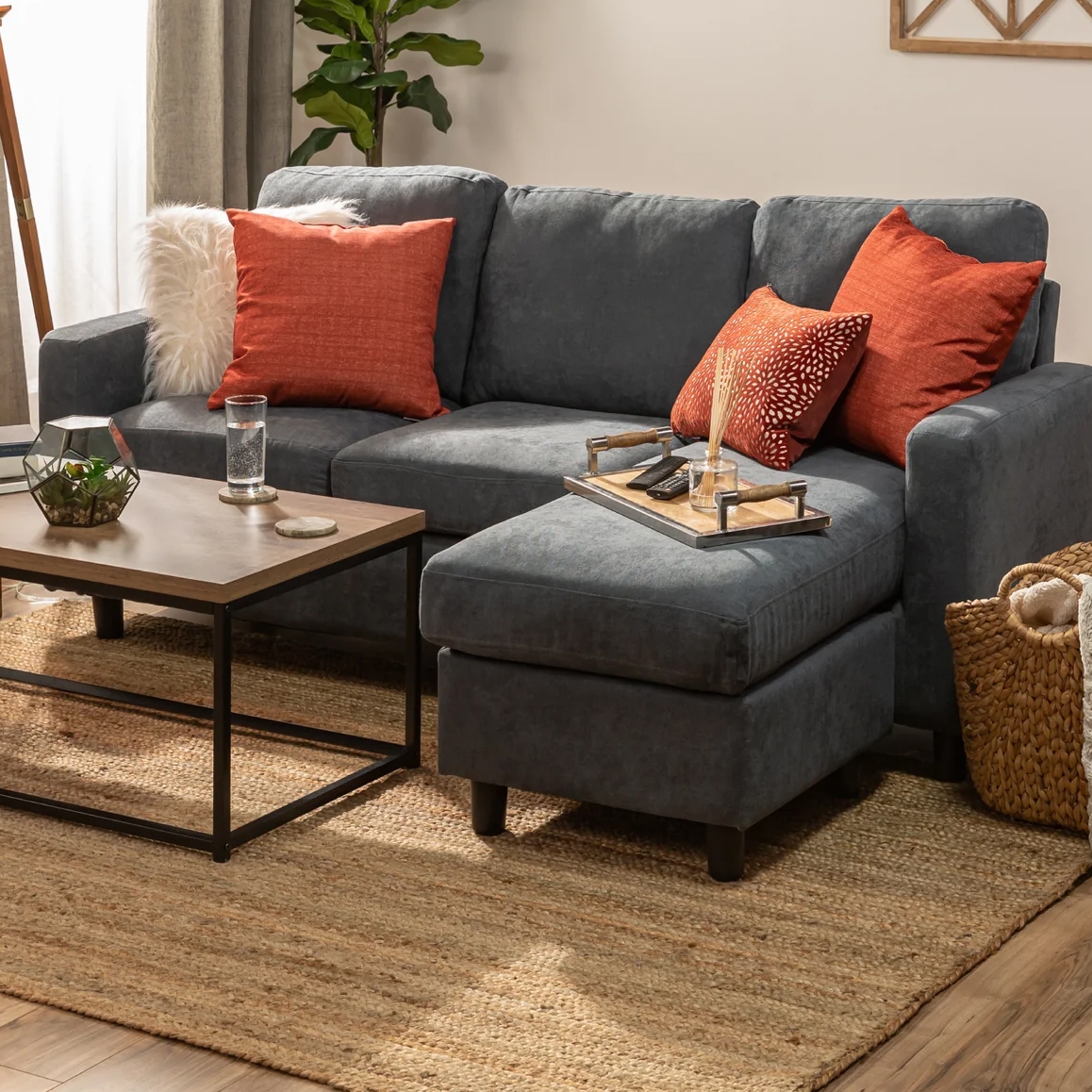 A dark gray sectional sofa with red-orange pillows is paired with a wooden and metal coffee table, featuring a glass of water and a plant. A woven basket and a natural fiber rug enhance the cozy living room ambiance.