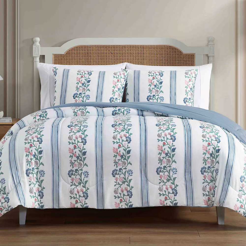 A bed with floral and striped patterned bedding, featuring a light blue and white color scheme. The headboard has a wicker design, while the room has a minimalist decor with light-colored walls and wooden flooring.
