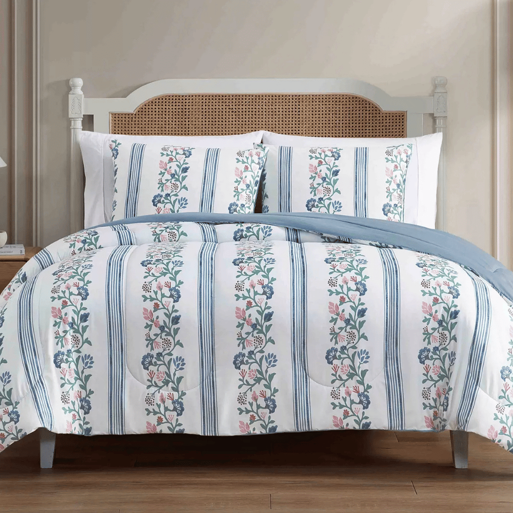 A bed with floral and striped patterned bedding, featuring a light blue and white color scheme. The headboard has a wicker design, while the room has a minimalist decor with light-colored walls and wooden flooring.
