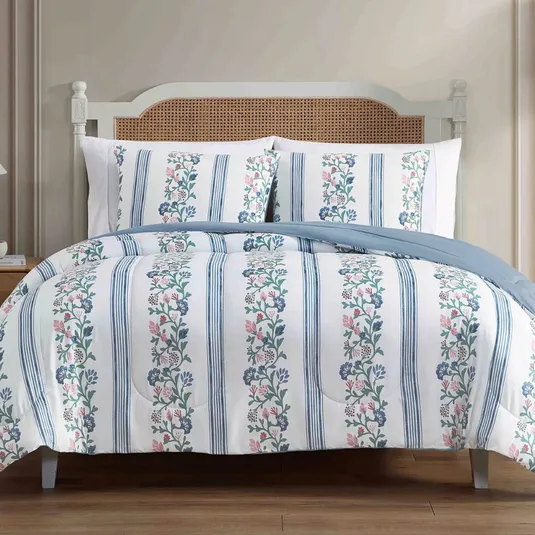 A bed with floral and striped patterned bedding, featuring a light blue and white color scheme. The headboard has a wicker design, while the room has a minimalist decor with light-colored walls and wooden flooring.