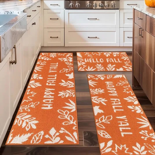 Three orange kitchen rugs with white leaf patterns and text reading \“HAPPY FALL Y'ALL\“ and \“HELLO FALL\“ placed on a wooden floor in a kitchen setting.
